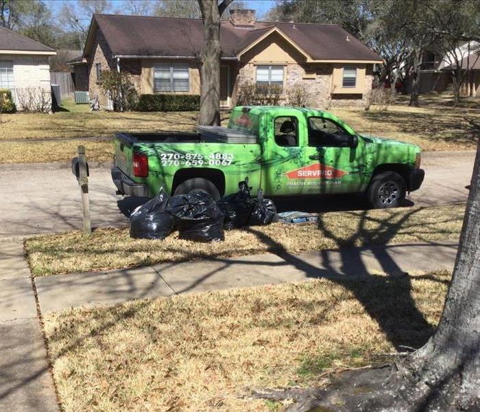 Green SERVPRO truck sitting in a neighborhood, parked on the curb with black bags of garbage sitting around